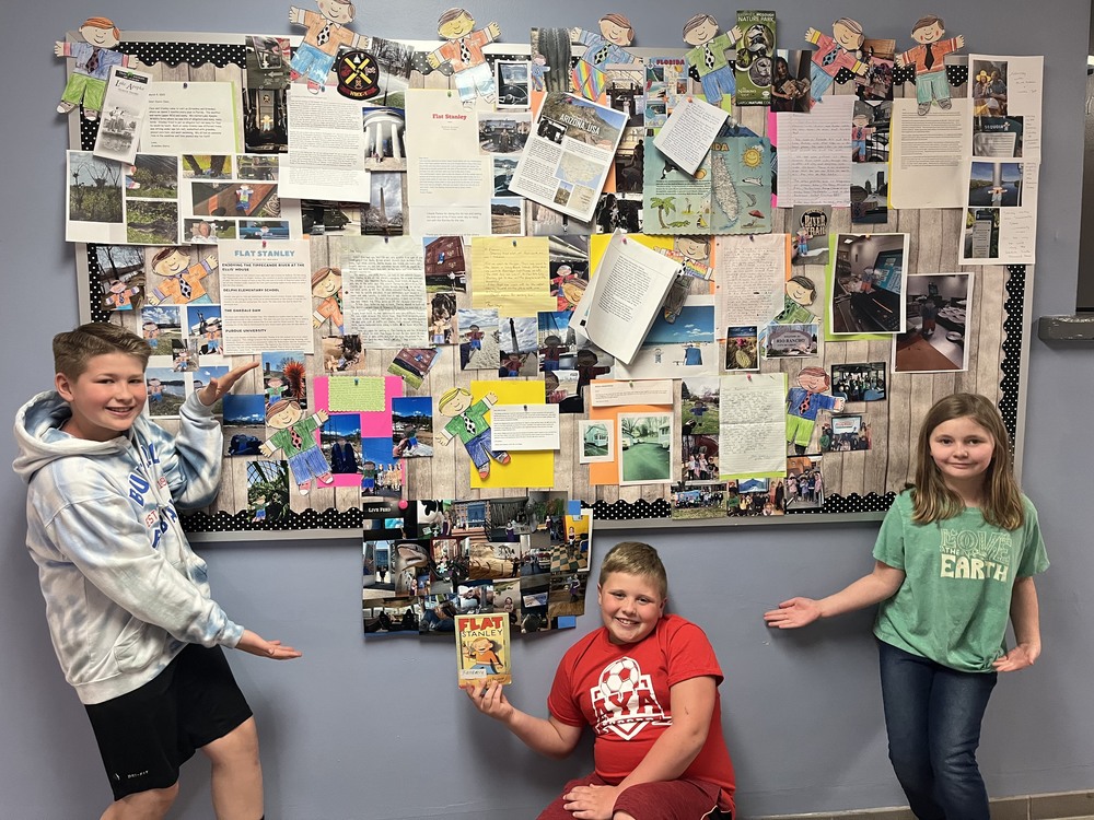 From left Levi Perry, Caden Morrison and Claira Deuel pose for a photo with the bulletin board chronicling the travels of Miss Lauren Kocher’s third graders’ Flat Stanleys. 