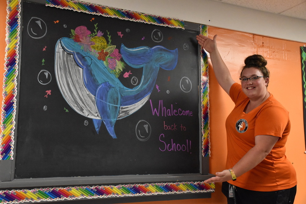 Custodian Brooke Schery poses for a photo next to her chalk drawing “whalecoming” elementary school students back for the 2022-23 school year. 