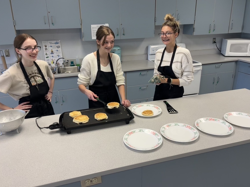 Three students in Mrs. Becky Sphar's “Foods on Your Own” class pose for a photo while preparing pancakes for a panel of judges.